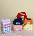 Five small fabric pouches shaped like fruits, each with a white label reading 'yams', displayed on a light pink surface against a beige wall. The pouches are red, navy blue, orange, pink, and pale yellow, with one green apple and one orange with leaf accents.
