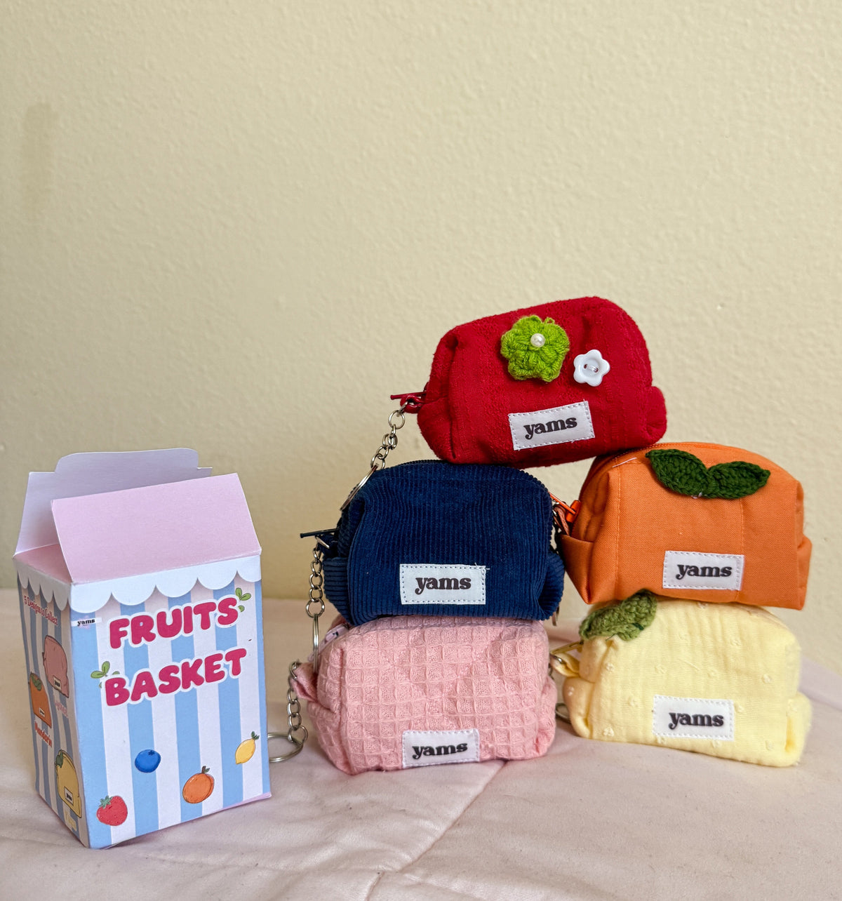 Five small fabric pouches shaped like fruits, each with a white label reading 'yams', displayed on a light pink surface against a beige wall. The pouches are red, navy blue, orange, pink, and pale yellow, with one green apple and one orange with leaf accents.